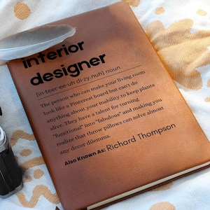 May include: A brown leather-bound journal with the definition of "interior designer" printed on the cover. The definition reads: "[in-teer-ee-uh di-zy.nuh] noun The person who can make your living room look like a Pinterest board but can't do anything about your inability to keep plants alive. They have a talent for turning "functional" into "fabulous" and making you realize that throw pillows can solve almost any decor dilemma. Also Known As: Richard Thompson"