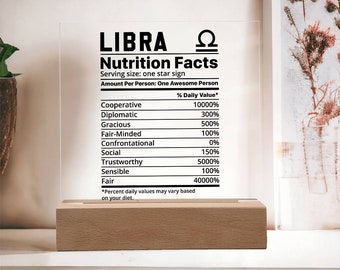 May include: A clear acrylic sign with the text "LIBRA Nutrition Facts" and the Libra astrological symbol. The sign lists various personality traits with a percentage value for each, such as "Cooperative 10000%", "Diplomatic 300%", and "Fair 40000%". The sign is sitting on a wooden base.