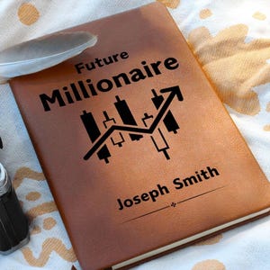 Peut inclure: Un journal en cuir marron avec les mots "Future Millionaire" et "Joseph Smith" imprimés sur la couverture. Un graphique boursier noir est également imprimé. Une plume blanche est posée sur le dessus.