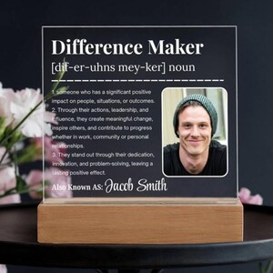Può includere: Una targa in acrilico trasparente con le parole "Difference Maker" e una definizione, insieme a una foto di una persona sorridente. La targa poggia su una base di legno e include il nome "Jacob Smith".