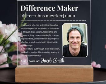 Pode incluir: Uma placa de acrílico transparente com as palavras "Difference Maker" e uma definição, juntamente com uma foto de uma pessoa sorrindo. A placa fica em uma base de madeira e inclui o nome "Jacob Smith".