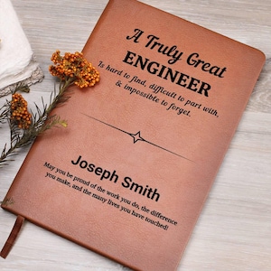 May include: A brown leather journal with the text "A Truly Great ENGINEER" and the name "Joseph Smith" printed on the cover. The journal includes a quote about engineers. A sprig of orange flowers rests on the side.