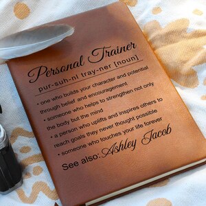 May include: A brown leather journal with the words "Personal Trainer" in elegant script. The definition of a personal trainer is printed below, along with the name "Ashley Jacob". A feather rests on the journal.