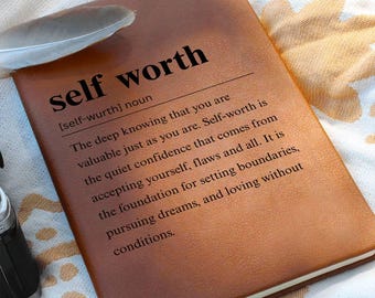 Puede incluir: Un diario de cuero marrón con las palabras "self worth" impresas en negro. La definición de autoestima está impresa debajo del título. Una pluma blanca descansa sobre el diario.