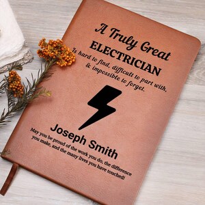 May include: A brown leather journal with the text "A Truly Great ELECTRICIAN" and a lightning bolt graphic. The journal includes the name "Joseph Smith" and a personalized message. A decorative sprig of orange flowers is placed near the journal.