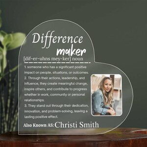 Puede incluir: Placa transparente de acrílico en forma de corazón con las palabras "Difference maker" y una definición. Incluye una foto de una mujer y el texto "Also Known As: Christi Smith".