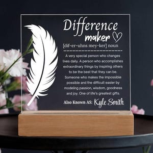May include: A clear acrylic plaque with the words "Difference Maker" and a definition, along with a feather graphic. The plaque sits on a wooden base and includes the name "Kyle Smith".
