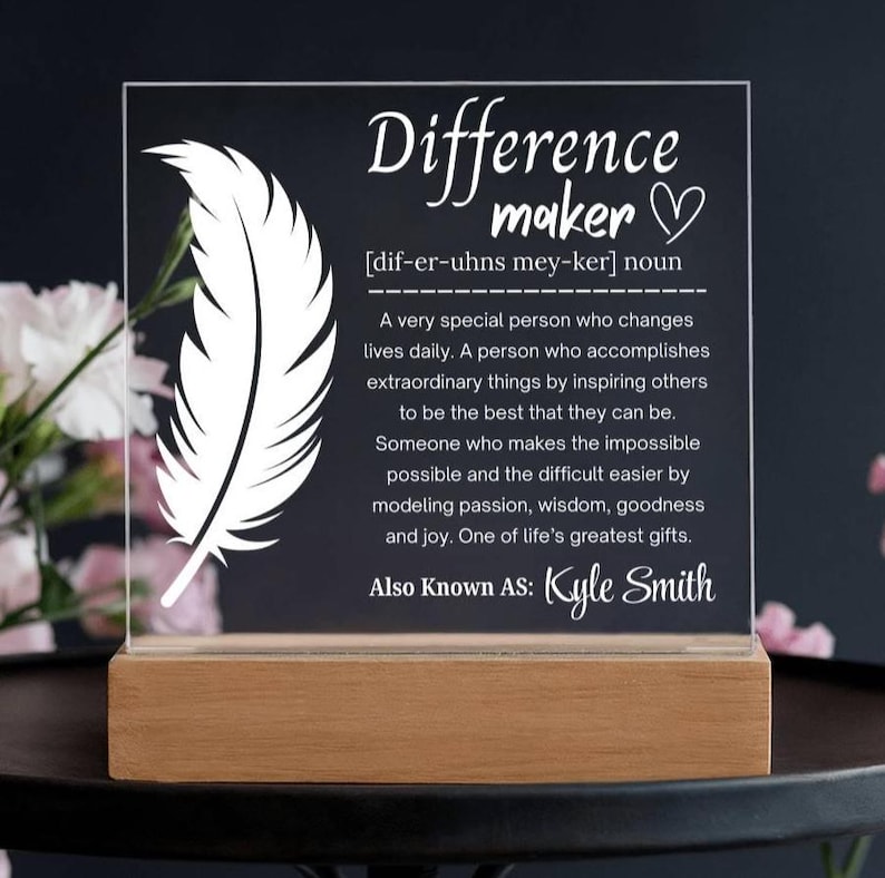 Op de afbeelding: Een heldere acrylplaat met de woorden "Difference maker" en een definitie, samen met een veergrafiek. De plaat staat op een houten voet en bevat de naam "Kyle Smith".