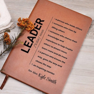 May include: A brown leather-bound notebook with the word "LEADER" printed in black. The notebook includes a definition of "leader" and the name "Kyle Smith". A sprig of orange flowers rests on the side.