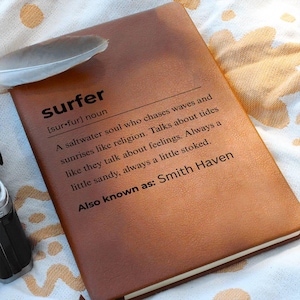 May include: A brown leather journal with the definition of "surfer" printed in black. The definition includes the phrase "a saltwater soul." A white feather rests on the journal. The text "Smith Haven" is also printed on the journal.