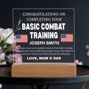 Op de afbeelding: Een heldere acrylplaat met de tekst "CONGRATULATIONS ON COMPLETING YOUR BASIC COMBAT TRAINING" en de naam "JOSEPH SMITH". Voorzien van twee Amerikaanse vlaggen en een houten voet. De tekst "LOVE, MOM & DAD" is ook zichtbaar.