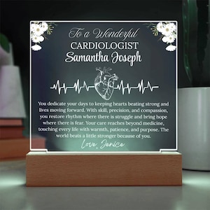 Puede incluir: Placa acrílica iluminada con base de madera, dedicada a una cardióloga llamada Samantha Joseph. La placa presenta una ilustración de corazón, una línea de latidos y un mensaje. Acentos florales blancos adornan las esquinas superiores.