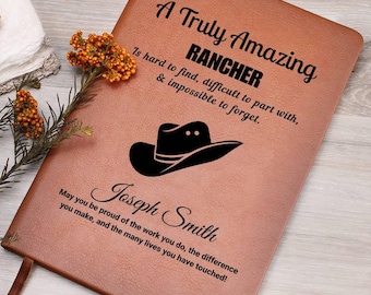 May include: A brown leather journal with the text "A Truly Amazing RANCHER" and a cowboy hat graphic. The journal includes the name "Joseph Smith" and a personalized message. A decorative flower and a ribbon bookmark are also visible.