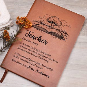 Puede incluir: Un diario de cuero marrón con una definición de la palabra "Teacher" y una ilustración de un libro y un árbol. El texto dice: "Teacher (n) [tee-cher] 1. A multi-tasking educational rockstar who lives to inspire and loves to encourage you to dream big 2. One who helps you gain knowledge, skills, experience and values Also Known as: Miss Robinson"