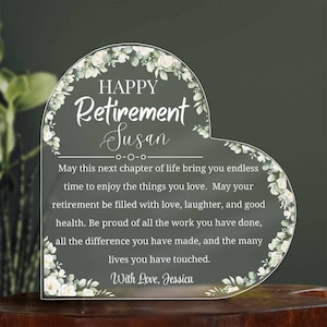 May include: A heart-shaped clear acrylic retirement plaque with floral accents. The plaque reads "HAPPY Retirement Susan" and includes a personalized message. The design features white flowers and green leaves, perfect for a retirement gift.