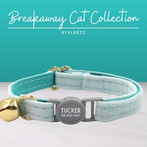 May include: A light blue breakaway cat collar with a gold bell and a gray tag that says "TUCKER 214-555-7647". The collar is made of soft, comfortable fabric and is designed to break away if caught on something.