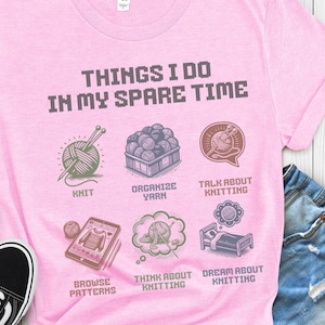 May include: Pink t-shirt with the text "THINGS I DO IN MY SPARE TIME" and illustrations of knitting related activities, including knitting, organizing yarn, talking about knitting, browsing patterns, thinking about knitting, and dreaming about knitting.