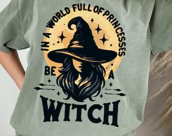 May include: A sage green t-shirt featuring a graphic print of a witch wearing a pointed hat against a yellow moon background. The text reads, "IN A WORLD FULL OF PRINCESSES BE A WITCH," with small star and broomstick illustrations surrounding the central figure.