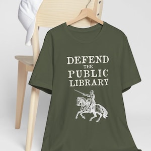 May include: A green t-shirt with the text "DEFEND THE PUBLIC LIBRARY" in white. A knight on horseback is depicted below the text.