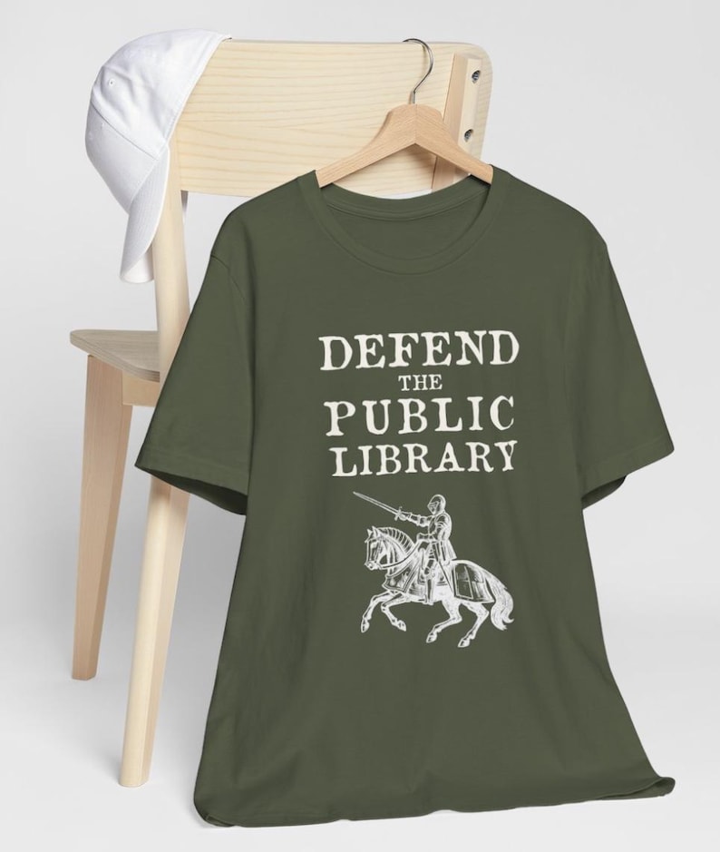 May include: A green t-shirt with the text "DEFEND THE PUBLIC LIBRARY" in white. A knight on horseback is depicted below the text.