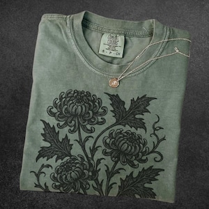 May include: Sage green t-shirt with a black floral design and a silver necklace. The shirt has a crew neck and the text "COMFORT COLORS" on the tag. The word "Sage" is at the bottom left.