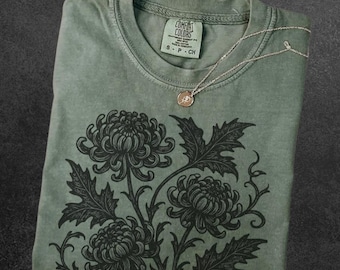 May include: Sage green t-shirt with a black floral design and a silver necklace. The shirt has a crew neck and the text "COMFORT COLORS" on the tag. The word "Sage" is at the bottom left.