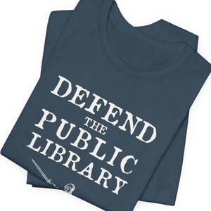 May include: A navy blue t-shirt with the text "DEFEND THE PUBLIC LIBRARY" in white. A sword and a knight's helmet are visible at the bottom of the shirt.