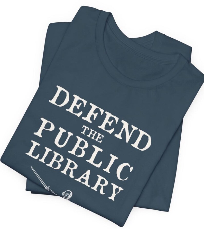 May include: A navy blue t-shirt with the text "DEFEND THE PUBLIC LIBRARY" in white. A sword and a knight's helmet are visible at the bottom of the shirt.