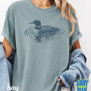 Puede incluir: Una camiseta de manga corta en color azul polvoriento con una ilustraci&oacute;n gr&aacute;fica en azul oscuro de un colimbo nadando en el agua. El ave se muestra de perfil con patrones de plumas detallados y ondas que rodean su cuerpo.