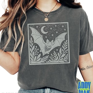 May include: A charcoal gray short-sleeve t-shirt featuring a square graphic print of a bat in flight, a crescent moon, stars, and foliage. The design is rendered in a light gray, ink-stamped style on the front center of the garment.