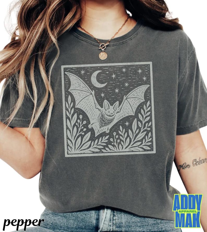 May include: A charcoal gray short-sleeve t-shirt featuring a square graphic print of a bat in flight, a crescent moon, stars, and foliage. The design is rendered in a light gray, ink-stamped style on the front center of the garment.
