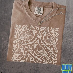 May include: Espresso-colored t-shirt with a white graphic of four birds perched on branches. The shirt has a crew neck and a tag that reads "Comfort Colors." A silver necklace with a small pendant rests on the shirt. The text "Espresso" is at the bottom.