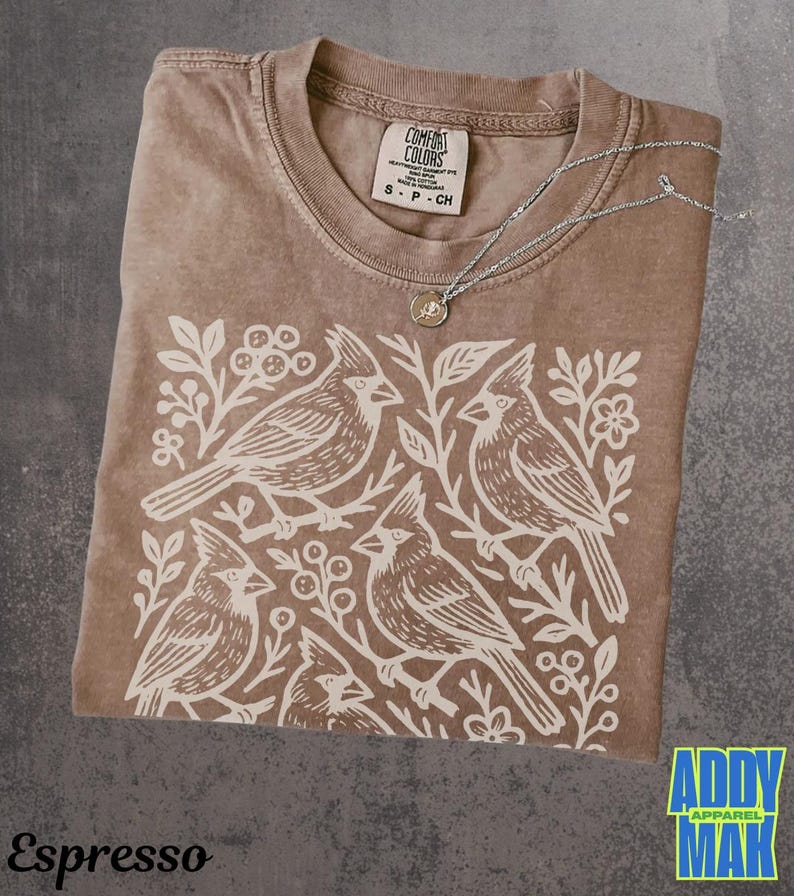 May include: Espresso-colored t-shirt with a white graphic of four birds perched on branches. The shirt has a crew neck and a tag that reads "Comfort Colors." A silver necklace with a small pendant rests on the shirt. The text "Espresso" is at the bottom.