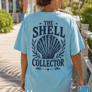 May include: A light blue short-sleeve t-shirt featuring a dark blue graphic on the back. The design includes a large seashell illustration centered between the text THE SHELL COLLECTOR, accented with small stars and leafy branch motifs.