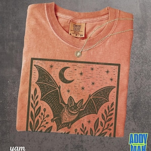 May include: A terracotta-colored t-shirt featuring a black graphic print of a bat in flight under a crescent moon and stars, framed by foliage. The shirt has a crew neckline with a Comfort Colors label and a thin gold-toned chain necklace.