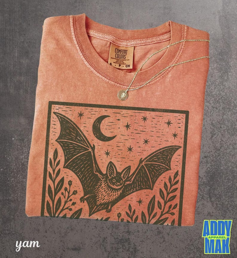 May include: A terracotta-colored t-shirt featuring a black graphic print of a bat in flight under a crescent moon and stars, framed by foliage. The shirt has a crew neckline with a Comfort Colors label and a thin gold-toned chain necklace.
