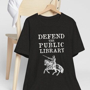 May include: Black t-shirt with white text that reads "DEFEND THE PUBLIC LIBRARY" and a white illustration of a knight on horseback.