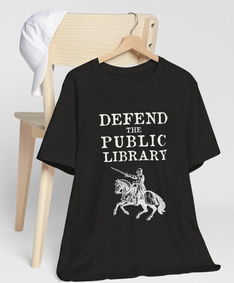 May include: Black t-shirt with white text that reads "DEFEND THE PUBLIC LIBRARY" and a white illustration of a knight on horseback.