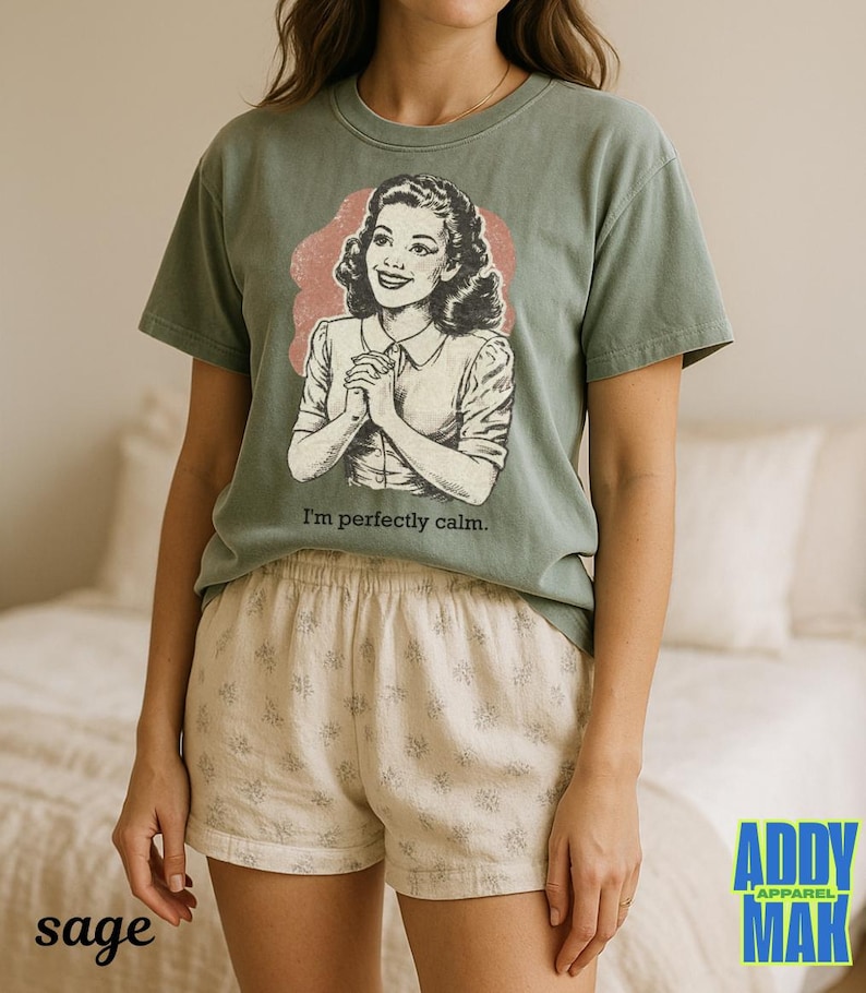 Op de afbeelding: Saliegroen t-shirt met een vintage illustratie van een vrouw en de tekst "I'm perfectly calm." De vrouw is afgebeeld met gevouwen handen. Het shirt wordt gecombineerd met cr&egrave;mekleurige shorts met een bloemenpatroon. Het merklogo staat rechtsonder.