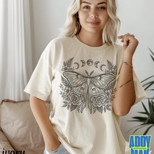 May include: Ivory-colored t-shirt featuring a detailed illustration of a moth with floral and moon phase elements. The graphic is in shades of gray. The shirt has short sleeves and a relaxed fit. The text "ADDY APPAREL MAK" is visible.