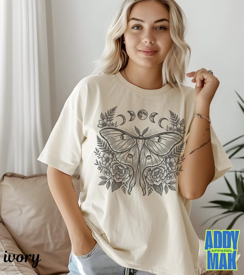 May include: Ivory-colored t-shirt featuring a detailed illustration of a moth with floral and moon phase elements. The graphic is in shades of gray. The shirt has short sleeves and a relaxed fit. The text "ADDY APPAREL MAK" is visible.