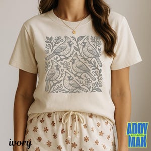 May include: Ivory-colored t-shirt featuring a gray cardinal bird design. The graphic includes five birds, branches, and floral elements. The text "ivory" is at the bottom left. The brand logo "ADDY MAK APPAREL" is at the bottom right.