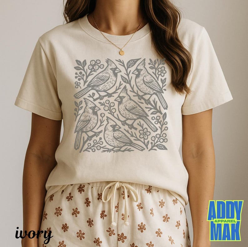 May include: Ivory-colored t-shirt featuring a gray cardinal bird design. The graphic includes five birds, branches, and floral elements. The text "ivory" is at the bottom left. The brand logo "ADDY MAK APPAREL" is at the bottom right.