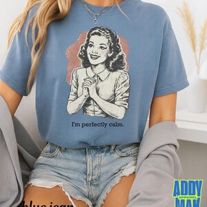 Op de afbeelding: Een blauw t-shirt met een vintage illustratie van een vrouw en de tekst "I'm perfectly calm." De afbeelding is in zwart-wit met een roodbruine achtergrond. Het shirt wordt gedragen met een spijkerbroekshort.