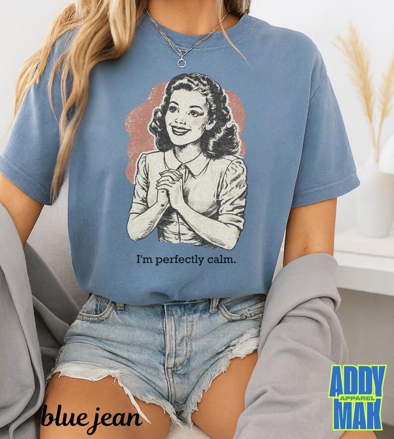 Op de afbeelding: Een blauw t-shirt met een vintage illustratie van een vrouw en de tekst "I'm perfectly calm." De afbeelding is in zwart-wit met een roodbruine achtergrond. Het shirt wordt gedragen met een spijkerbroekshort.