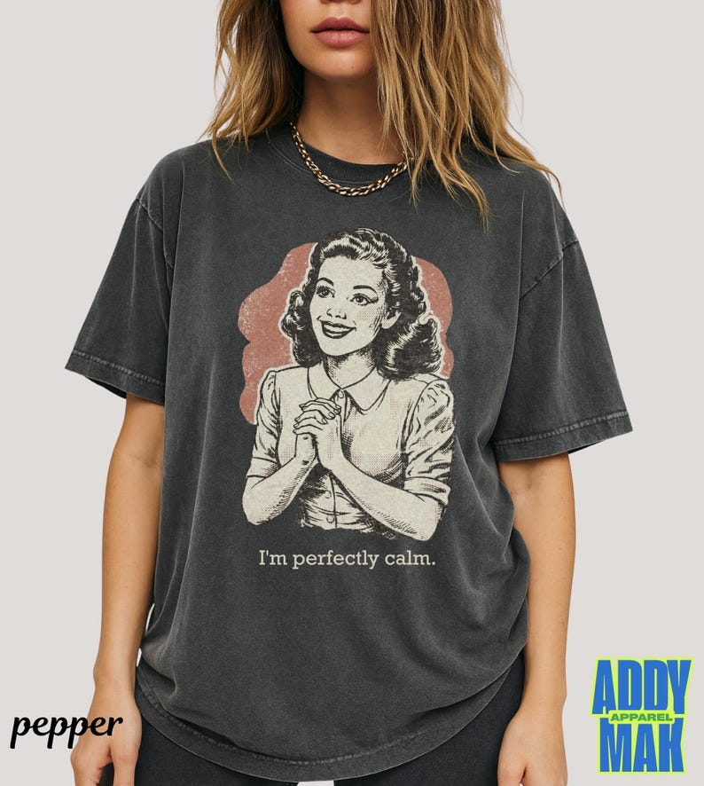 Op de afbeelding: Donkergrijs T-shirt met een vintage illustratie van een vrouw en de tekst "I'm perfectly calm." De afbeelding is in retrostijl, met een vrouw in een overhemd met kraag en een roze-rode achtergrond. Het shirt heeft een relaxte pasvorm.
