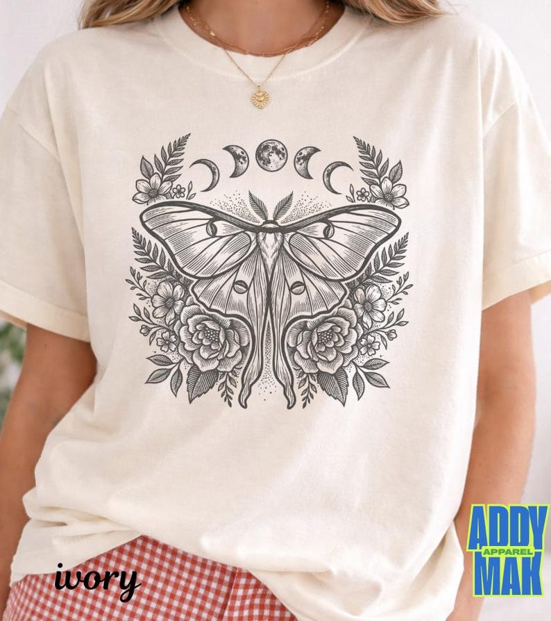 May include: Ivory-colored t-shirt featuring a detailed black and white illustration of a moth with floral and moon phase elements. The text "ivory" is printed at the bottom left. The brand logo "ADDY MAK" is at the bottom right.