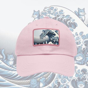 May include: A light pink baseball cap featuring a rectangular patch with a blue and white wave design and cat illustrations. The cap has a curved brim and a fabric strap closure.