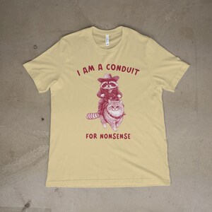 May include: A yellow t-shirt with a red graphic of a raccoon wearing a cowboy hat riding a cat. The text "I am a conduit for nonsense" is printed on the shirt.