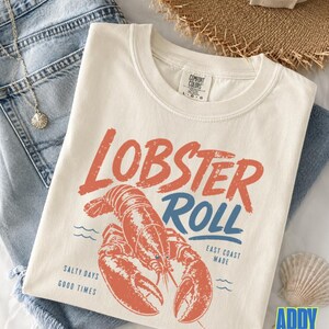 Pu&ograve; includere: T-shirt color avorio con grafica arancione invecchiata con la scritta "Lobster Roll" sopra un'illustrazione dettagliata di un'aragosta. La maglietta presenta anche il testo "East Coast Made" e "Salty Days Good Times".